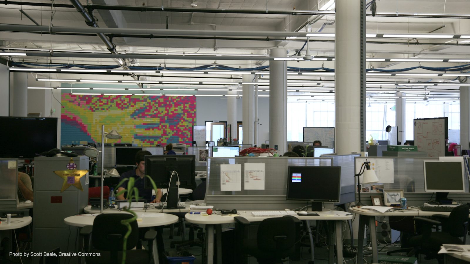 A wide photo of an open-plan office, with desks, monitors and a wall covered in colorful sticky notes.