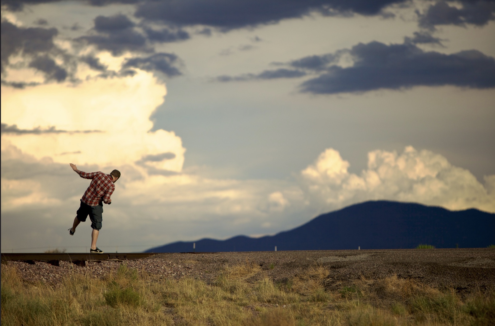 A small person balances beside a railway line beneath a vast dramatic sky and distant mountains