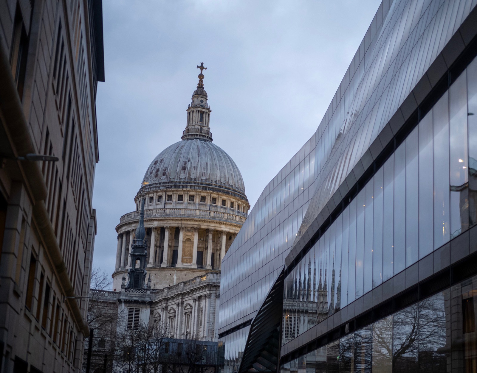 A large stone dome framed between a dark building and a curved glass facade beneath a pale grey sky