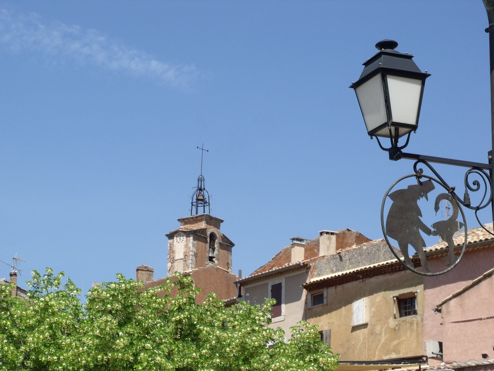 Roussillon’s bell tower, rooftops and an old wrought-iron shop sign against blue sky