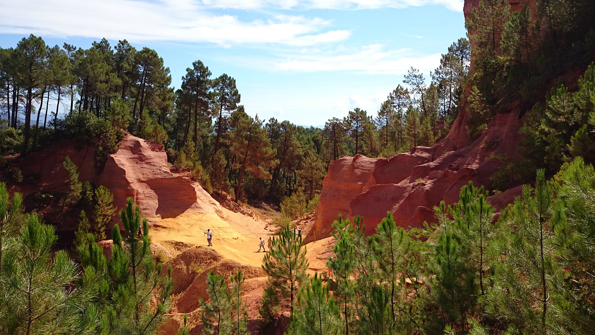 Visitors walking below a vast red ochre quarry face and pine trees