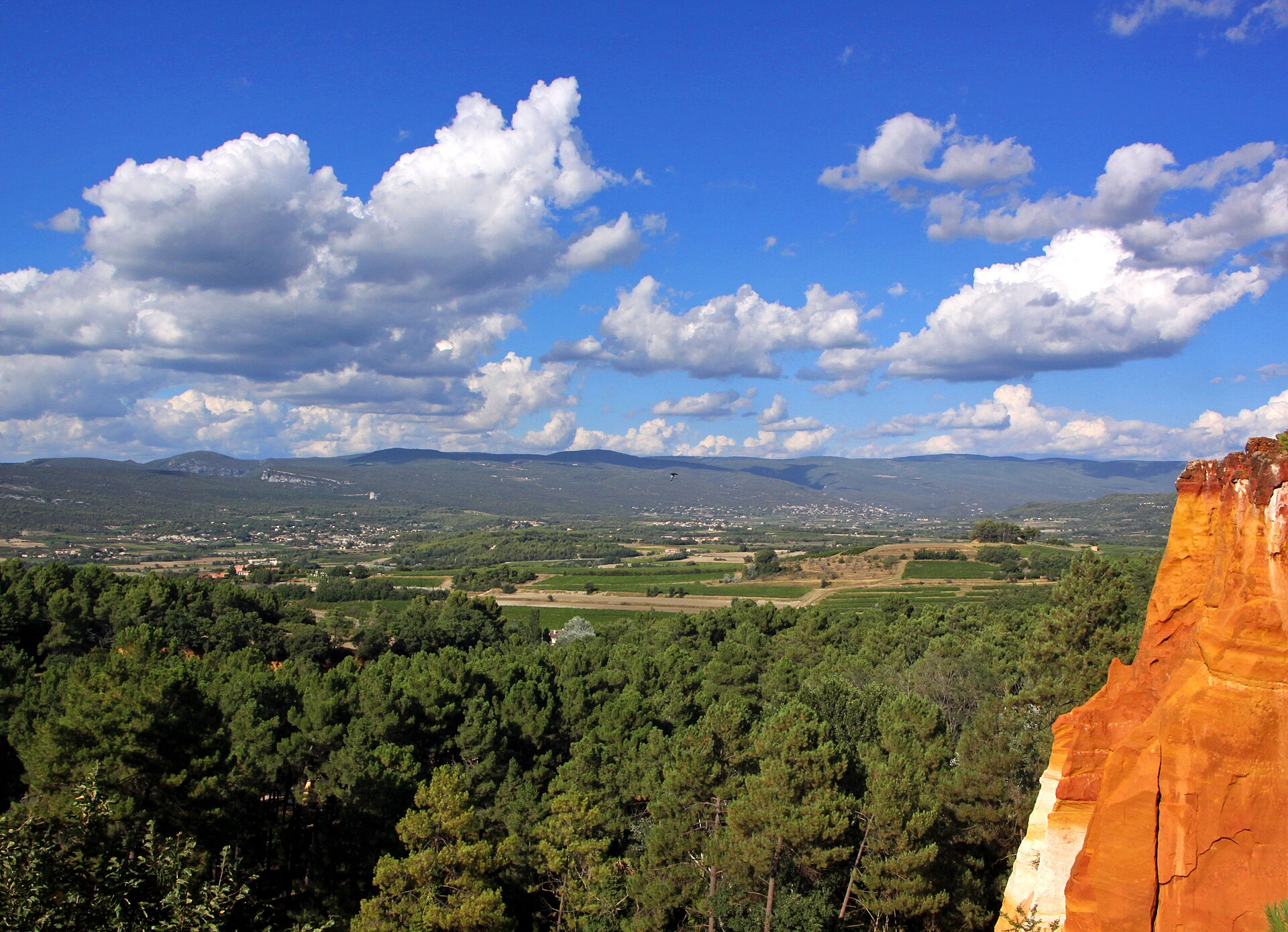 An orange ochre cliff rising between green fields and distant blue hills
